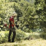 Honda UMK450 XE 47CC Brushcutter in action with a man in safety gear using it to tidy unkempt undergrowth in a green, wooded area.